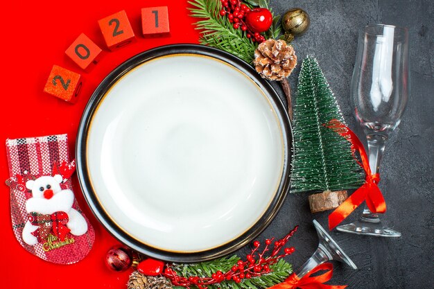 Vista aérea de platos de cena, accesorios de decoración, ramas de abeto, números de calcetines navideños en una servilleta roja y copa de vidrio de árbol de Navidad sobre fondo oscuro