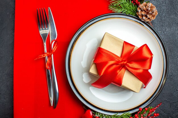 Vista aérea de platos de cena con regalo y ramas de abeto cubiertos accesorios de decoración cono de coníferas en una servilleta roja