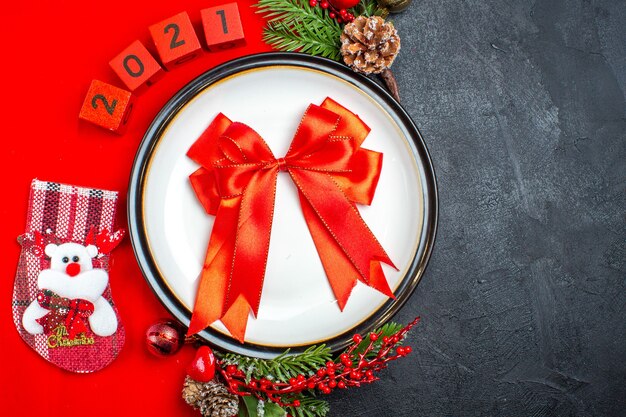 Vista aérea del regalo con cinta en el plato de cena, accesorios de decoración, ramas de abeto y números, calcetín de navidad en una servilleta roja sobre un fondo negro