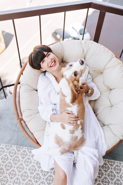Vista aérea de la risa joven con peinado corto y perro beagle mirando hacia arriba con interés. Retrato de niña alegre en vestido blanco acostado en silla redonda con lindo perrito.