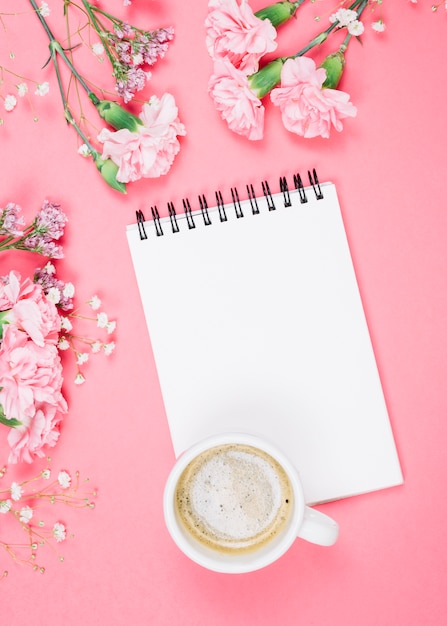 Una vista aérea de la taza de café en la libreta en blanco con claveles; gypsophila; flores de limonium sobre fondo rosa