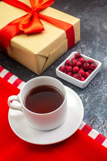 Vista aérea de una taza de té negro sobre una toalla roja y galletas en un plato blanco de regalo con cinta roja y cornel sobre superficie oscura