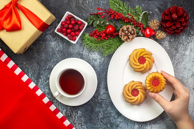 Vista aérea de una taza de té negro, una toalla roja y una mano tomando galletas de un plato blanco, regalo de accesorios de año nuevo con cornel de cinta roja sobre una superficie oscura.