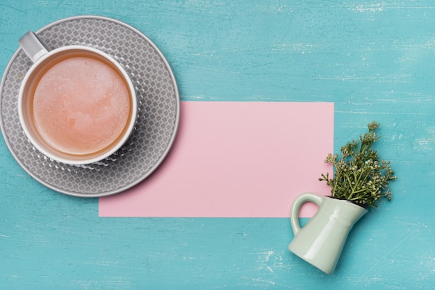 Foto gratuita una vista aérea de la taza de té con el papel y el florero rosados ​​en blanco en el contexto azul de la textura