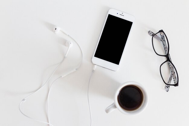 Vista aérea del teléfono móvil con auriculares conectados, taza de café y anteojos