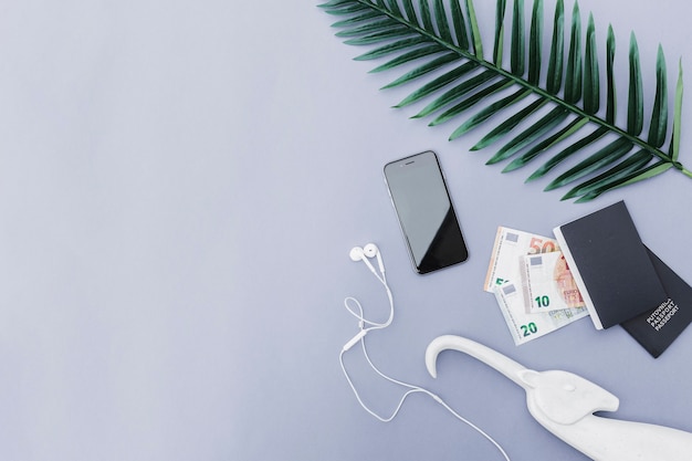 Vista aérea del teléfono móvil con auriculares, moneda euro, pasaporte y hojas sobre fondo gris