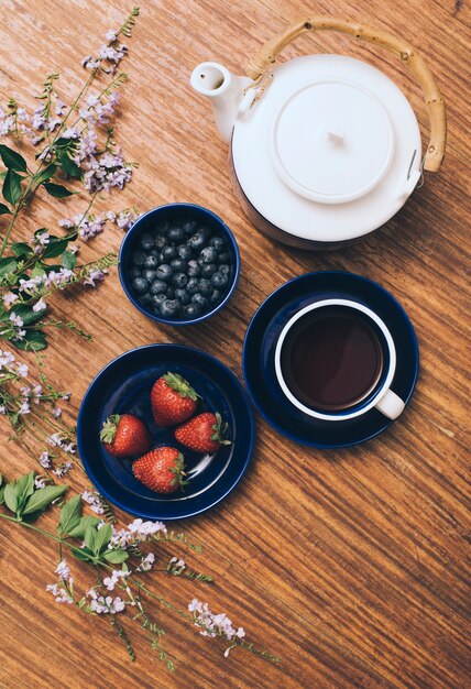 Una vista aérea de la tetera; arándanos; taza de café; y fresa con flores sobre fondo de madera.