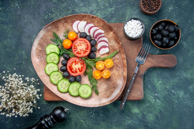 Vista aérea de verduras frescas picadas en una placa marrón sobre tabla de cortar de madera aceitunas en un tazón flor de ajos de sal sobre fondo de colores mezclados