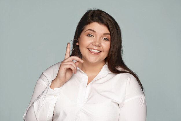 Vista aislada del gerente gordito regordete de mujer joven con aretes redondos y camisa blanca formal mordiendo la lengua y levantando el dedo, teniendo una gran idea, mirando con expresión feliz