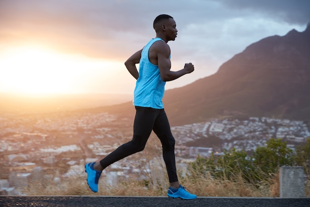 La vista al aire libre del corredor masculino joven activo cubre un largo destino en la mañana al amanecer, corre sobre la vista de las montañas, tiene bíceps, está vestido con ropa deportiva, respira profundamente, disfruta del clima de verano.