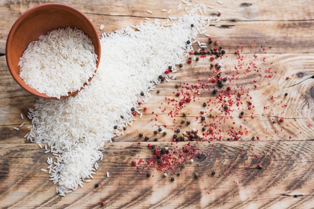 Vista de ángulo alto de arroz orgánico en un tazón y especias de pimientos negros sobre papel tapiz de madera