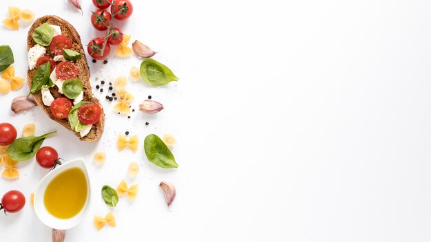 Vista de ángulo alto de bruschetta con pasta cruda farfalle; diente de ajo; tomate; petróleo; Hoja de albahaca contra aislados sobre fondo blanco