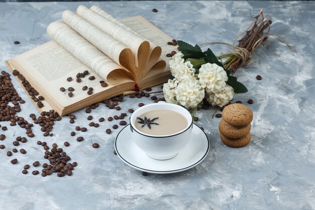 Vista de ángulo alto de café en taza con galletas, granos de café, flores, libro sobre fondo de yeso gris. horizontal