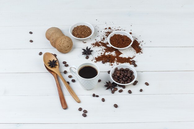 Vista de ángulo alto de café en taza con granos de café, café molido, especias, galletas, cucharas de madera sobre fondo de madera. horizontal