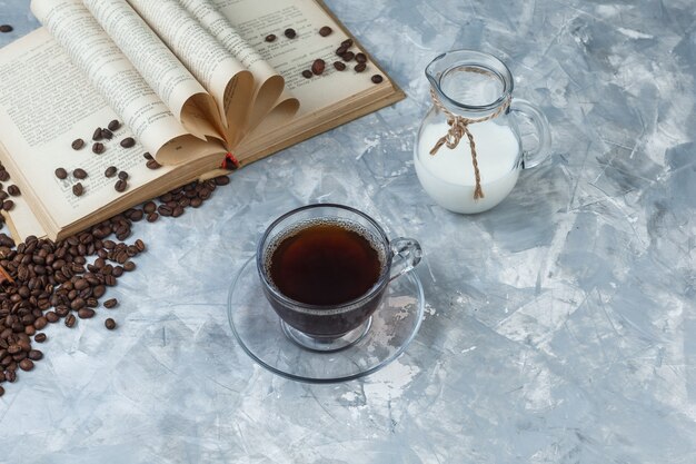 Vista de ángulo alto de café en taza con granos de café, libro, leche sobre fondo gris sucio. horizontal