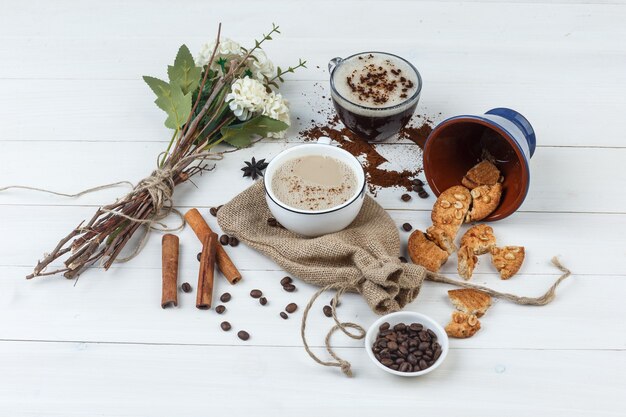 Vista de ángulo alto de café en tazas con granos de café, galletas, flores, canela sobre fondo de madera y saco.