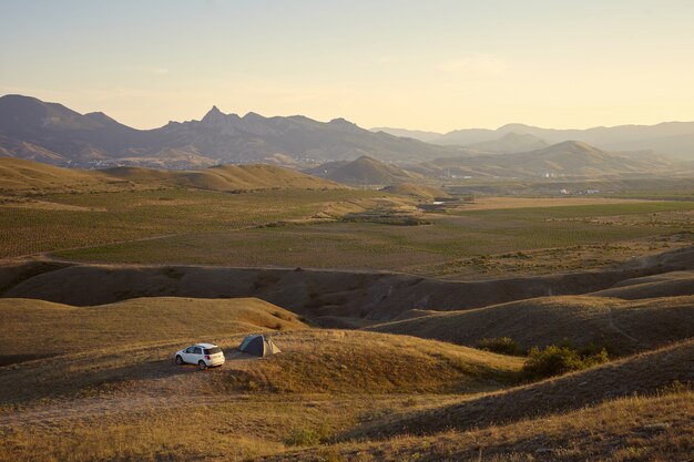 Vista de ángulo alto del camping en el pintoresco paisaje de las montañas. Turistas acampando en una zona montañosa con carpa y coche blanco aparcado junto a él. Concepto de naturaleza, turismo, viajes, vacaciones y senderismo.