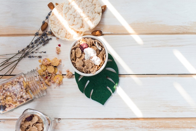 Foto gratuita vista de ángulo alto de copos de maíz en un tazón cerca de granola derramada y galletas de arroz en superficie de madera