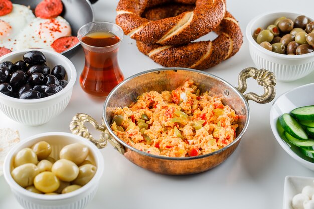 Vista de ángulo alto deliciosas comidas en sartén y olla con ensalada, encurtidos, una taza de té, panecillos turcos sobre superficie blanca