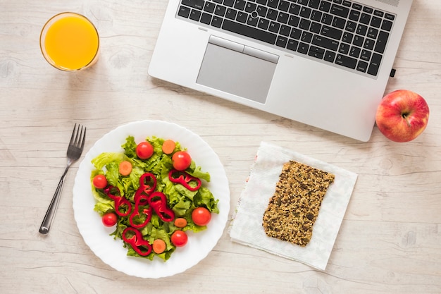 Foto gratuita vista de ángulo alto de ensalada; jugo; barra de granola; frutas con laptop en mesa