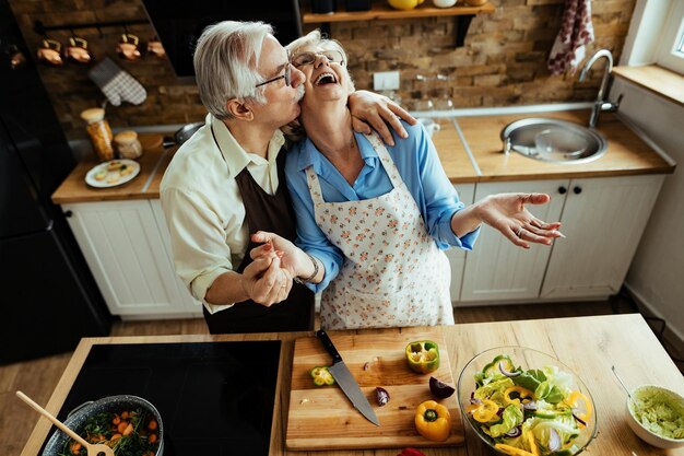 Vista de ángulo alto de la feliz pareja madura divirtiéndose mientras prepara la comida en la cocina. El hombre está besando a su esposa.