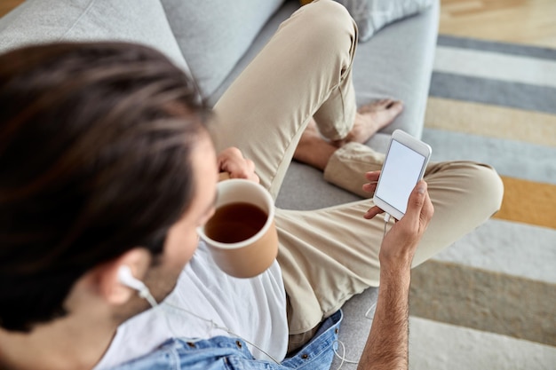Vista de ángulo alto de un hombre tomando café y usando un teléfono inteligente mientras se relaja en el sofá Copiar espacio