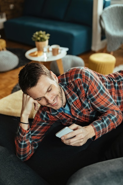 Vista de ángulo alto del joven feliz usando teléfono celular y mensajes de texto mientras está acostado en el sofá