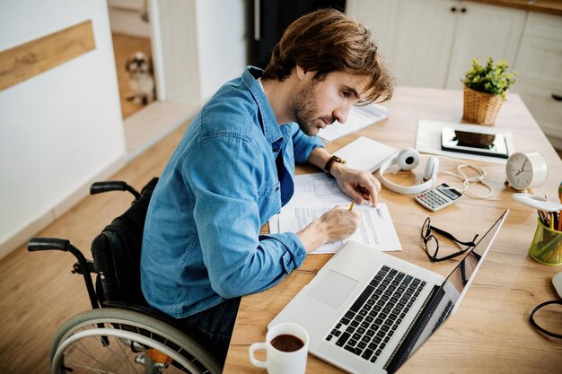 Vista de ángulo alto del joven hombre de negocios en silla de ruedas haciendo papeleo mientras trabaja en la computadora portátil en casa