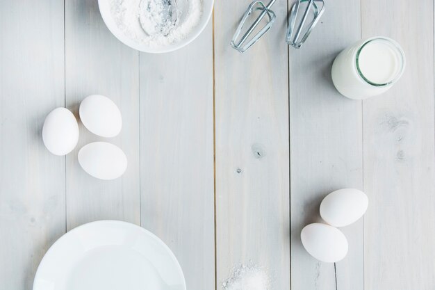 Vista de ángulo alto de la leche; harina; Batidor y huevos sobre tabla de madera.