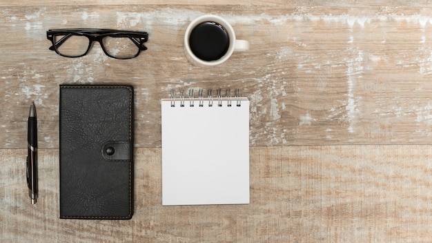 Vista de ángulo alto de la libreta en blanco; diario; bolígrafo; taza de café; anteojos en escritorio de madera