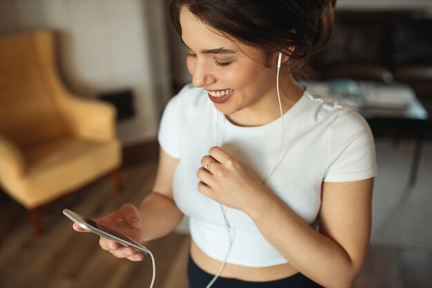 Vista de ángulo alto de linda adolescente alegre en mensajes superiores ajustados blancos con amigos o escuchando nuevas pistas de música usando la aplicación en línea en el teléfono móvil