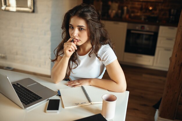 Vista de ángulo alto de maestra joven concentrada sentada en la mesa con computadora portátil, sosteniendo la pluma con expresión facial pensativa, preparándose para la lección en línea, escritura a mano en el cuaderno