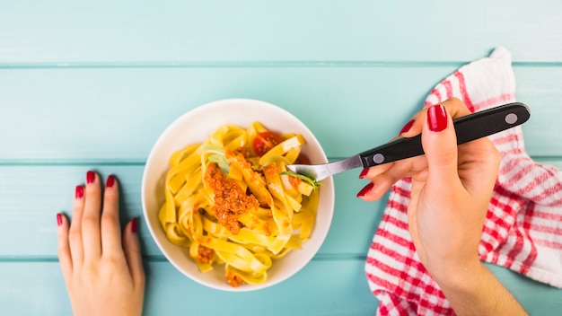 Foto gratuita vista de ángulo alto de la mano de una mujer comiendo pasta de tagliatelle en el escritorio