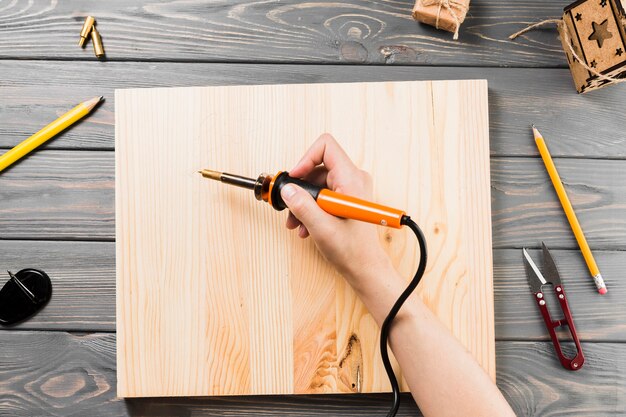 Vista de ángulo alto de la mano que sujeta la máquina de soldar en el tablero de madera para cortar la forma