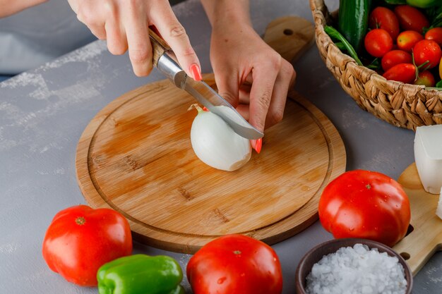 Vista de ángulo alto mujer cortando cebolla en la mitad en la tabla de cortar con cuchillo, pimiento verde, pepino, sal en superficie gris