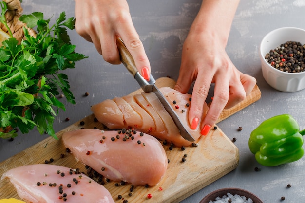 Vista de ángulo alto mujer cortando pechuga de pollo en la tabla de cortar con pimiento, pimiento verde sobre superficie gris