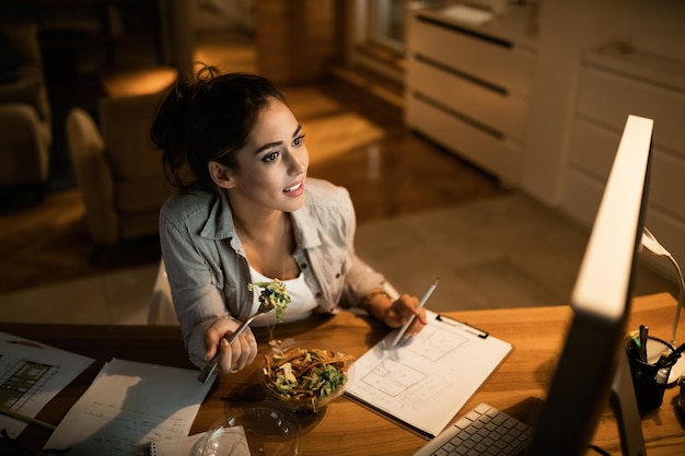 Vista de ángulo alto de mujer joven comiendo ensalada saludable mientras trabaja en PC de escritorio por la noche en casa