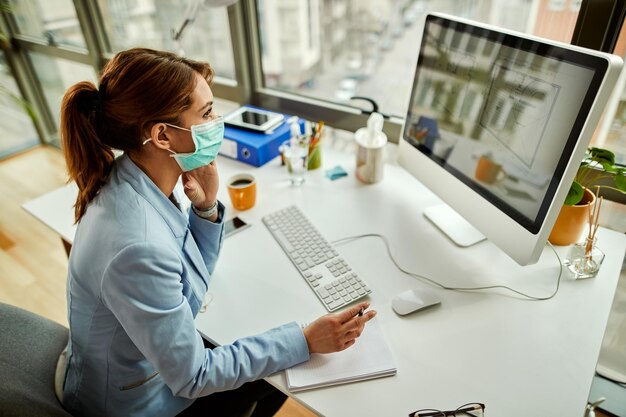 Vista de ángulo alto de la mujer de negocios con máscara facial analizando planos en PC de escritorio mientras trabaja en la oficina