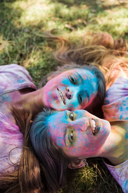 Vista de ángulo alto de mujeres jóvenes con color holi en la cara que yace en el césped mirando la cámara