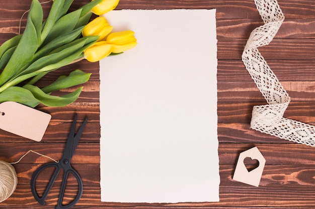 Vista de ángulo alto de papel en blanco; Flores amarillas; cortar con tijeras; cuerda; Forma de corazón y encaje sobre fondo de madera