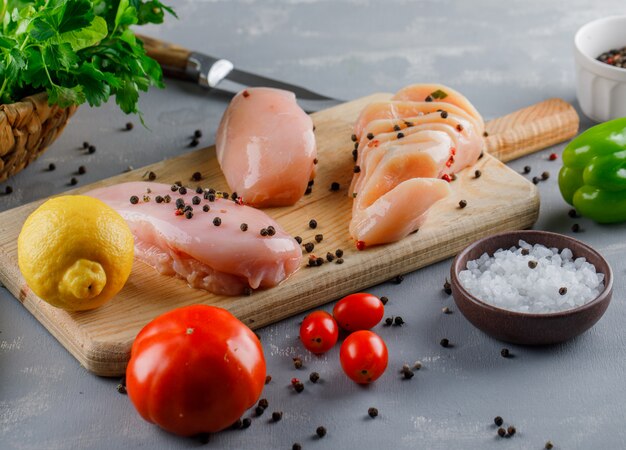 Vista de ángulo alto pechugas de pollo en tabla de cortar con limón, tomate, sal en superficie gris