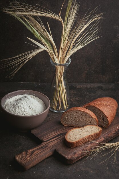 Vista de ángulo alto rebanada de pan con harina en un tazón y trigo en la tabla de cortar y marrón oscuro.