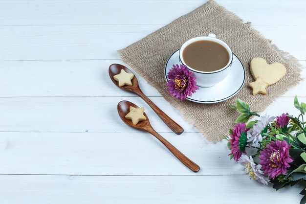 Vista de ángulo alto taza de café, galletas en forma de corazón y estrella en un pedazo de saco con flores, galletas en cucharas de madera sobre fondo de tablero de madera blanca. horizontal