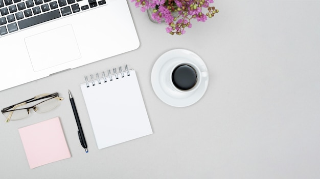 Vista de ángulo alto de la taza de café; ordenador portátil; lente; Bloc de notas espiral en maceta en mesa gris