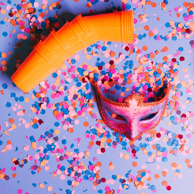 Vista de ángulo alto de vasos de plástico y máscara de fiesta elegante con confeti sobre fondo azul