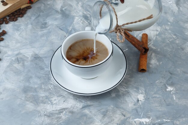 Vista de ángulo alto vertiendo leche en una taza de café con granos de café, ramas de canela sobre fondo gris sucio. horizontal