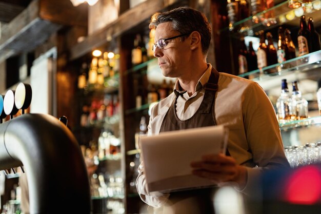 Vista de ángulo bajo del barman revisando la lista de inventario mientras trabaja en un pub.