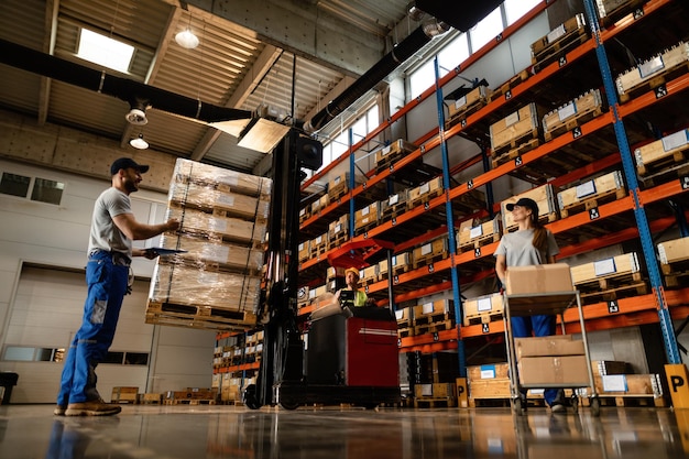 Vista de ángulo bajo de los felices trabajadores del almacén que se comunican mientras trabajan con el envío en el compartimento de almacenamiento industrial