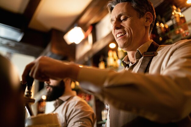 Vista de ángulo bajo de feliz barista vertiendo cerveza de barril mientras trabaja en un pub.