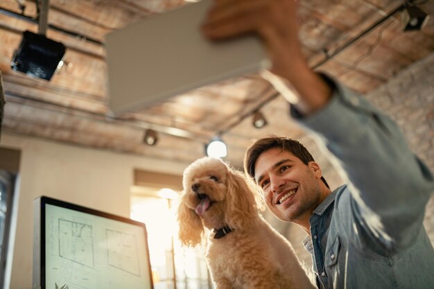 Vista de ángulo bajo de feliz freelancer divirtiéndose mientras toma selfie con un perro en la oficina